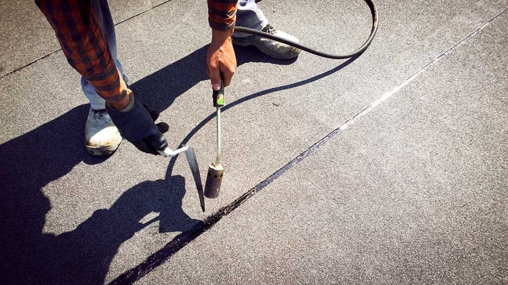 A person in work clothes using a tarp to cover roof damage after a storm, with ladders and scattered debris visible.