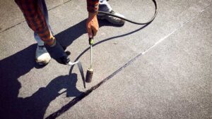 A person in work clothes using a tarp to cover roof damage after a storm, with ladders and scattered debris visible.