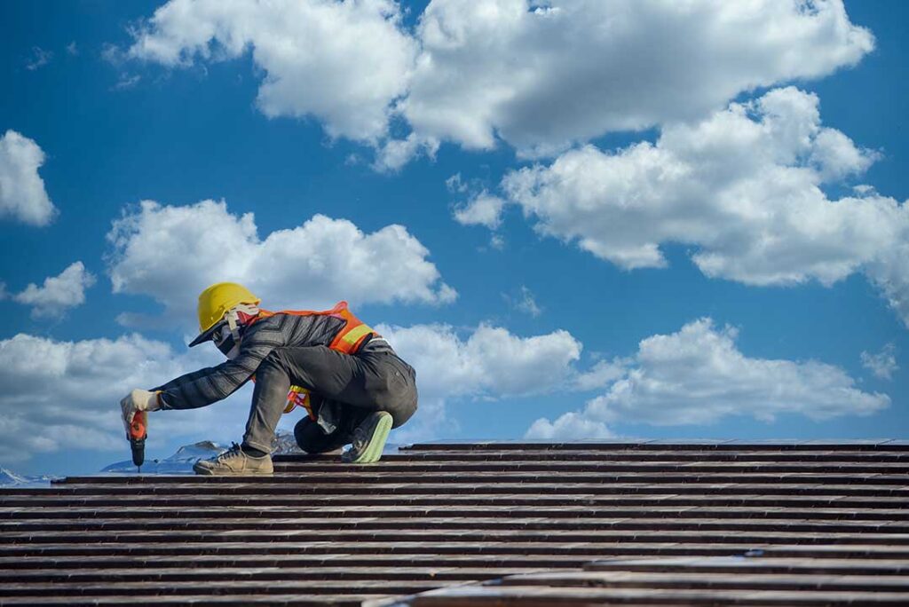 Person clearing leaves from a roof gutter beside asphalt shingles and a downspout.